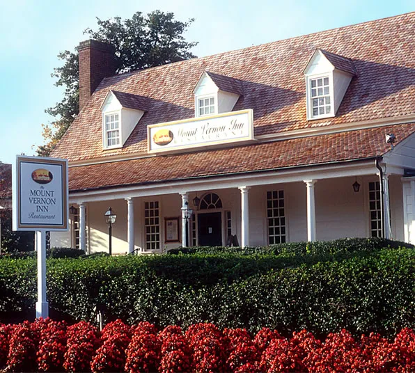 A charming white building with a shingled roof houses the Mount Vernon Inn Restaurant, surrounded by green bushes and red flowers under a clear blue sky. A sign in front reads Mount Vernon Inn Restaurant.