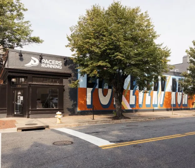 A street view of Pacers Running store with a colorful mural that says “For Every Run” on the side wall, bordered by trees and an empty sidewalk on a sunny day.