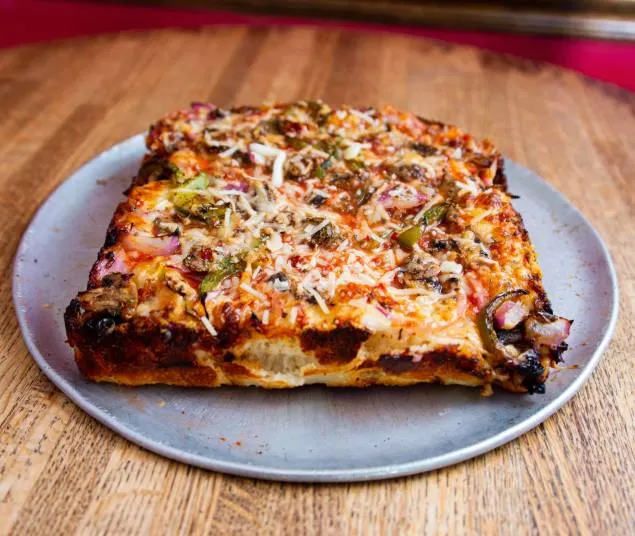 A square slice of Detroit-style pizza topped with cheese, vegetables, and herbs is served on a round metal plate atop a wooden table.