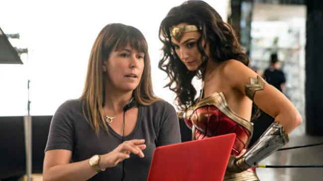 A woman wearing a headset speaks and gestures toward a red laptop, while another woman dressed as Wonder Woman stands beside her, listening intently.