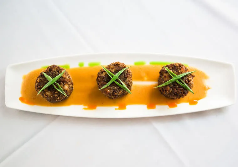 Three round crab cakes topped with green garnish are arranged in a row on a rectangular white plate, served over a layer of orange sauce on a white tablecloth background.