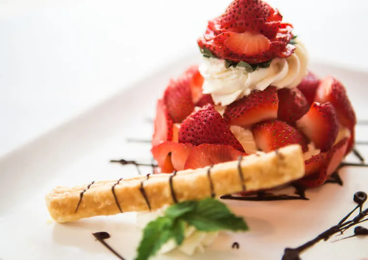 A dessert topped with fresh sliced strawberries and whipped cream, garnished with a pastry stick drizzled in chocolate and a sprig of mint, served on a white plate with chocolate sauce.