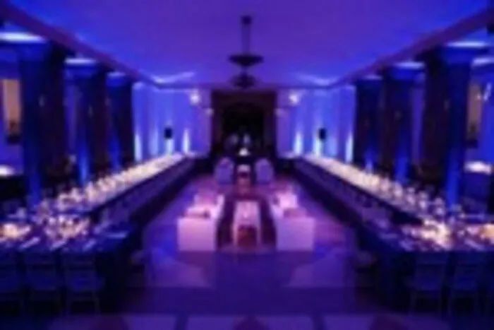 A large banquet hall is illuminated with blue and purple lights, featuring two long dining tables set for an event, with decorative place settings and candles, and lounge seating in the center.