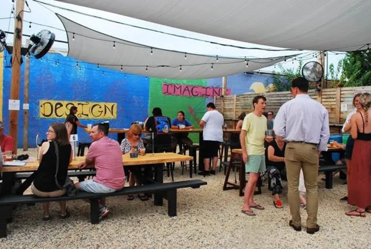 People sit and stand in an outdoor patio area with wooden tables under a canopy. Colorful murals on the walls say DESIGN and IMAGINE. Some people are eating, drinking, and talking in a relaxed, social atmosphere.