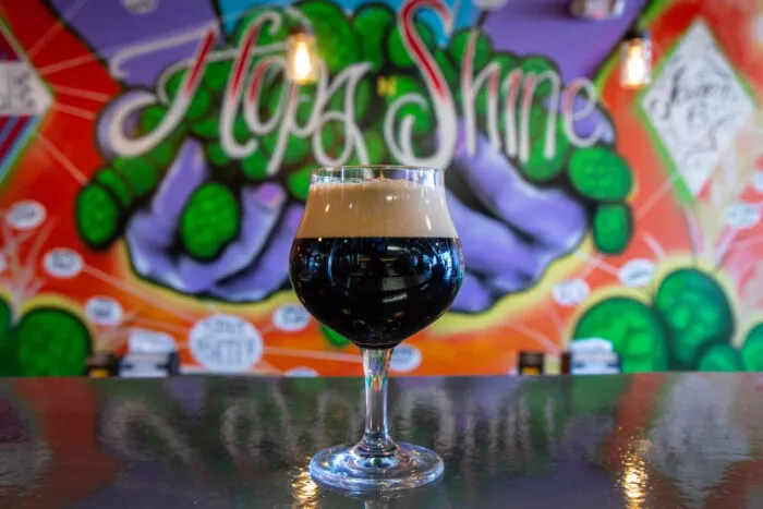 A glass of dark beer with a thick, creamy foam head sits on a reflective surface, with a colorful mural featuring the words Hop Shine and green hops blurred in the background.