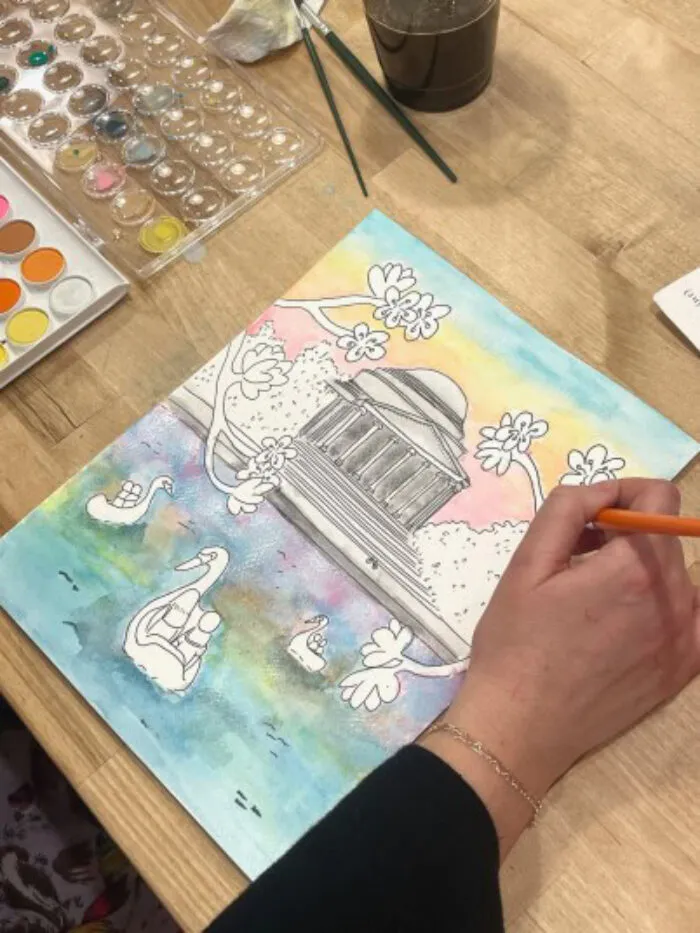 A person is painting a watercolor scene featuring a domed building, swans on a pond, and flowers. Paints, brushes, and a cup of water are on the wooden table beside the artwork.