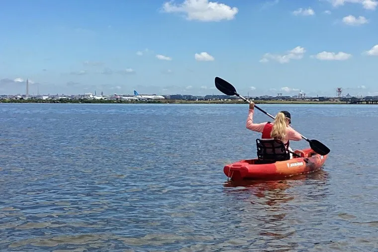 A person with long blond hair paddles a red kayak on calm water under a blue sky, with an airport and airplanes visible in the background.