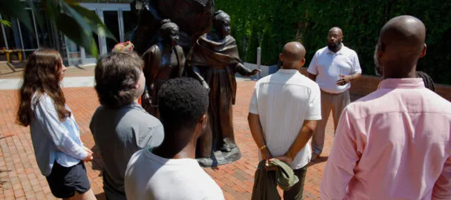 A group of people stands in a semi-circle around a historical statue outdoors, listening to a man in a white shirt speaking. The setting is sunny with greenery and a building in the background.