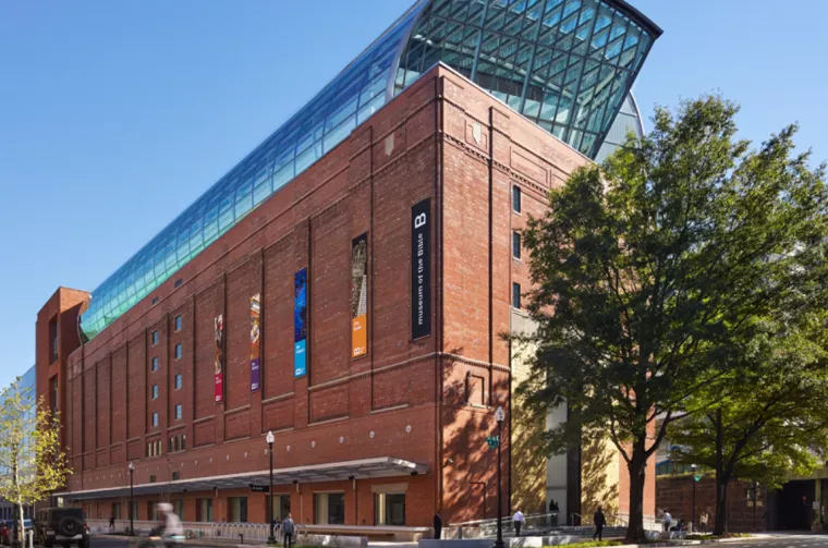 A large red-brick building with tall banners, large windows, and a modern glass rooftop structure; trees line the sidewalk on a sunny day.