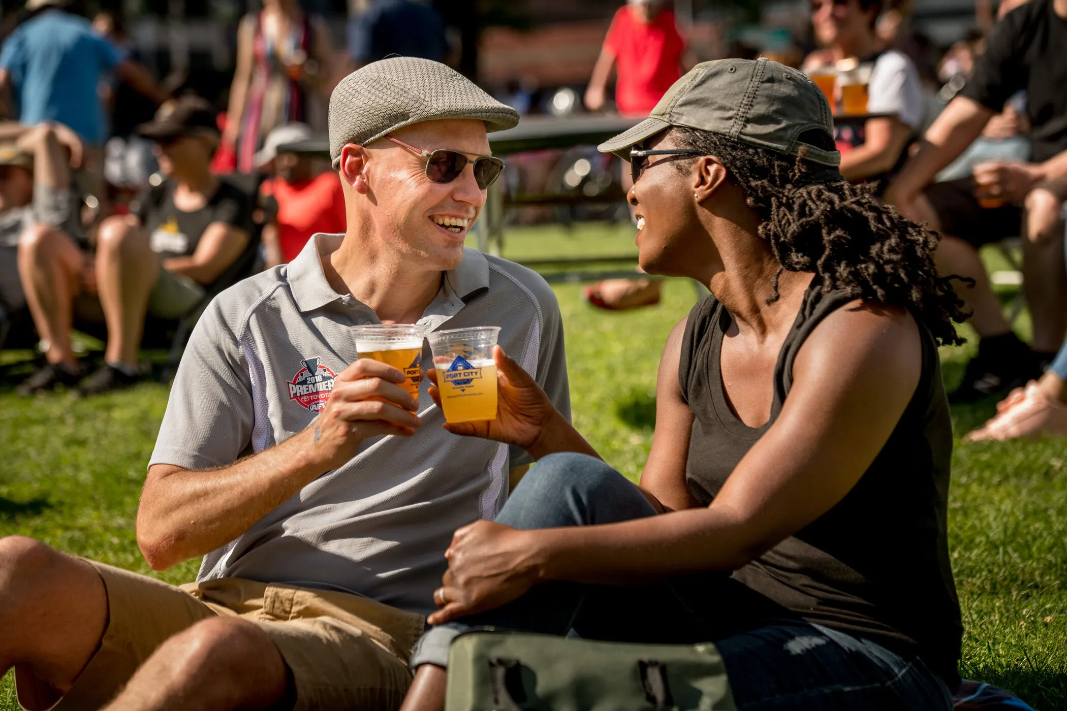 A man and woman sit on grass at an outdoor event, smiling and clinking plastic cups of beer—a perfect example of fun things to do this summer. People are sitting and walking in the background on a sunny day.