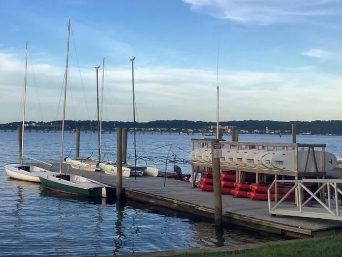 A wooden dock with sailboats tied alongside and stacks of kayaks and red paddleboards. Calm blue water and a distant shoreline with buildings and trees are visible under a partly cloudy sky.