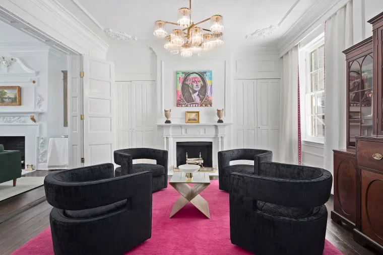 A modern living room with four black chairs around a white coffee table, a bright pink rug, a white fireplace with art above it, and a chandelier hanging from a white ceiling.