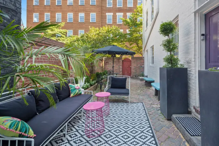 A stylish outdoor patio with black-and-white furniture, a patterned rug, pink tables, potted plants, and a black umbrella. Brick walls and a large building are visible in the background.