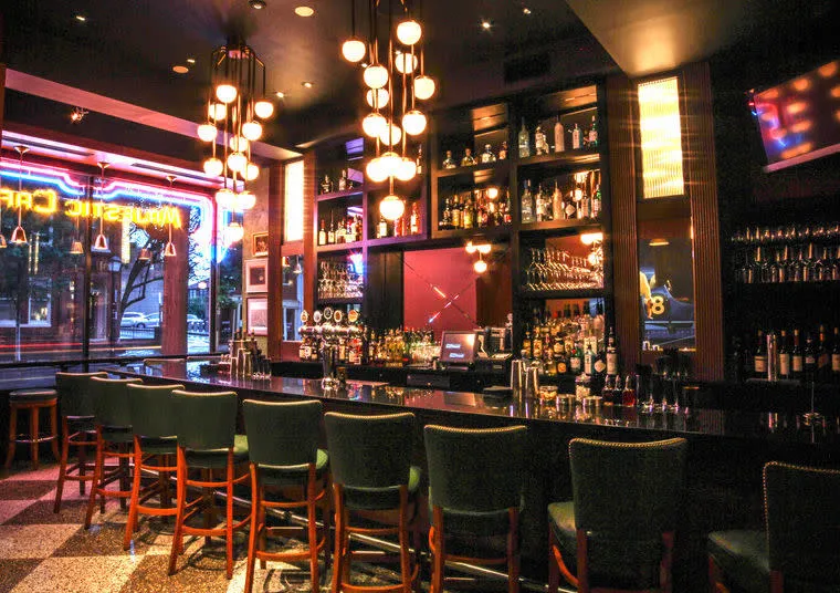 A stylish bar interior with a long dark counter, green barstools, hanging globe lights, shelves stocked with bottles, and a large window featuring neon signs. The atmosphere is warm and inviting.