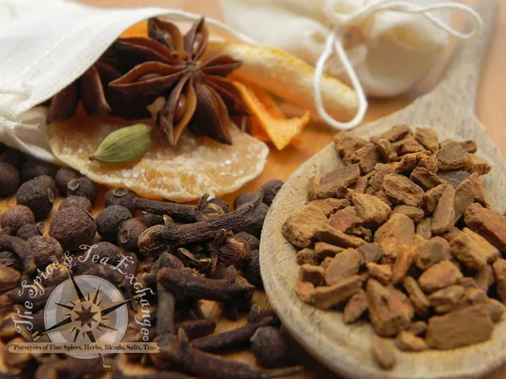 A close-up of assorted spices including cinnamon bark on a wooden spoon, dried cloves, star anise, cardamom, and rock sugar, with a white cloth pouch in the background.
