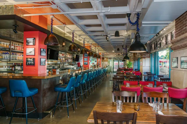 Bright restaurant interior with a long bar lined with blue stools, wooden tables with mismatched chairs, and colorful decor. Large windows let in natural light, creating an inviting atmosphere.