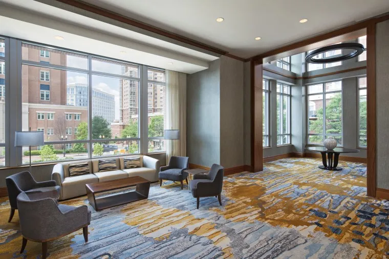 Modern lounge area with large windows, stylish chairs, a sofa, lamps, and a coffee table. Patterned carpet covers the floor, and city buildings and trees are visible outside the bright, spacious room.