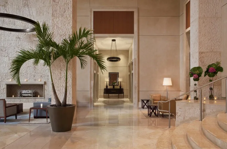 A spacious, elegant hotel lobby with marble floors, a large potted palm, modern seating, decorative lighting, and a hallway leading to a table with a framed mirror and flowers.