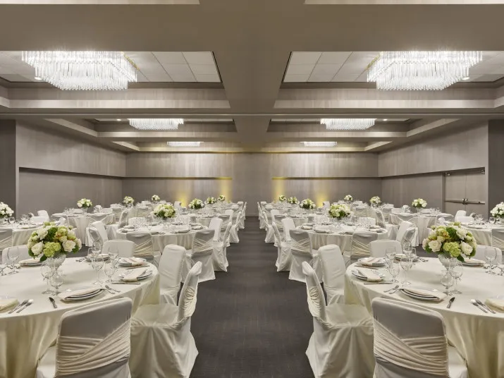 Elegant banquet hall with round tables covered in white cloths, set for a formal event. Each table has a centerpiece of white flowers. The room features modern chandeliers and neutral-toned walls.