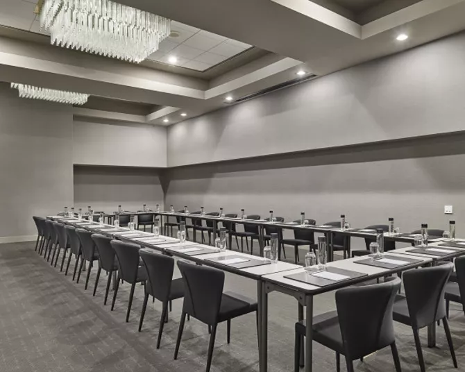 A modern conference room with gray walls and carpet, set up in a U-shape with black chairs and tables. Each place has a notepad, pen, glass, and bottle of water. A large chandelier hangs from the ceiling.