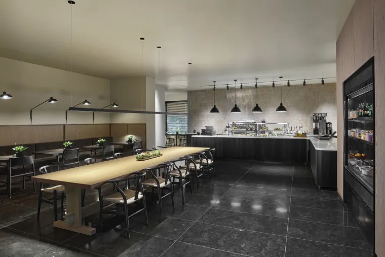 Modern dining area with long wooden tables and black chairs, indoor plants as centerpieces, black tile flooring, pendant lights, and a sleek counter with shelves holding snacks and drinks. Large windows provide natural light.