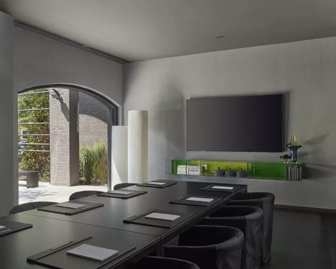 Modern conference room with a long table, black chairs, notepads, large wall-mounted TV, and shelves with decor. Natural light enters through a large arched window showing greenery outside.