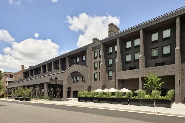 A modern, three-story hotel building with dark brick exterior, large windows, and an arched entrance; outdoor seating with white umbrellas is visible along the sidewalk under a partly cloudy sky.