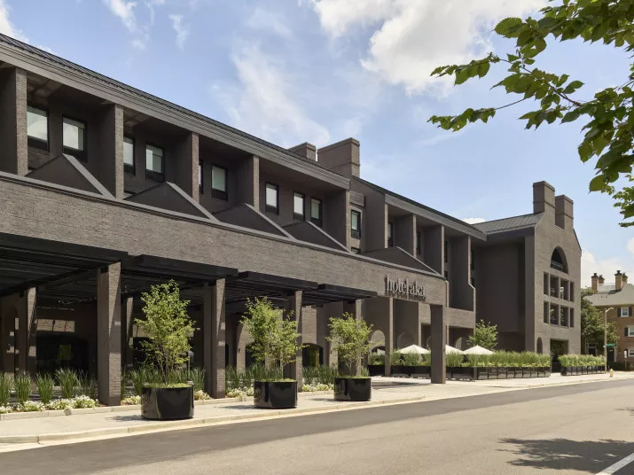 A modern, three-story brick building with large windows and a covered entryway, with Hotel Chelsea signage. Young trees and large planters line the sidewalk under a partly cloudy sky.