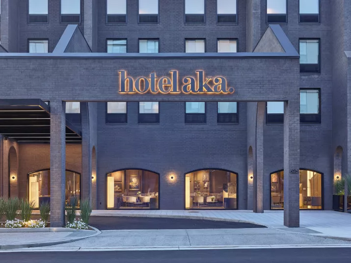 The exterior of a modern hotel building features large windows and a prominent sign reading hotelaka in glowing letters above the entrance. The facade is made of dark bricks.