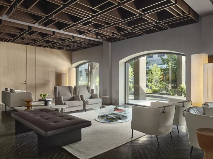 Modern living room with large windows, neutral-toned furniture, a black geometric ceiling, a glass coffee table, and a white rug. Natural light fills the space, and greenery is visible outside.