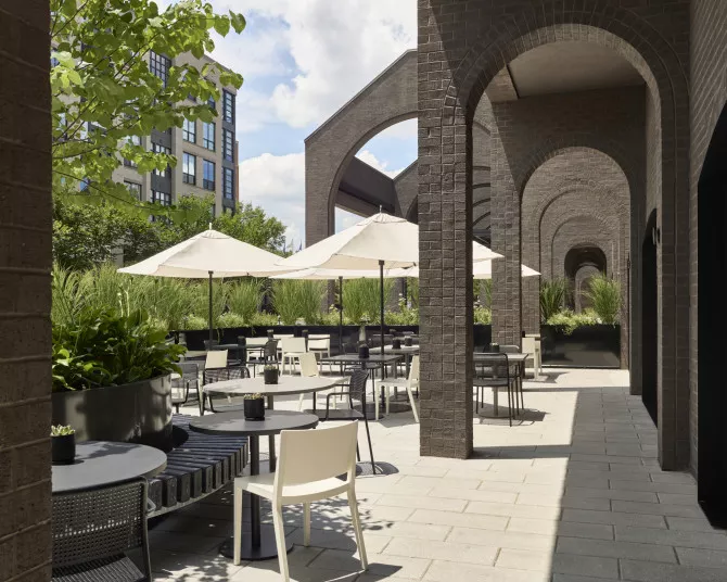 Modern outdoor patio with black brick arches, round tables, white chairs, and white umbrellas. Lush green plants line the seating area. A nearby building and partly cloudy sky are visible in the background.