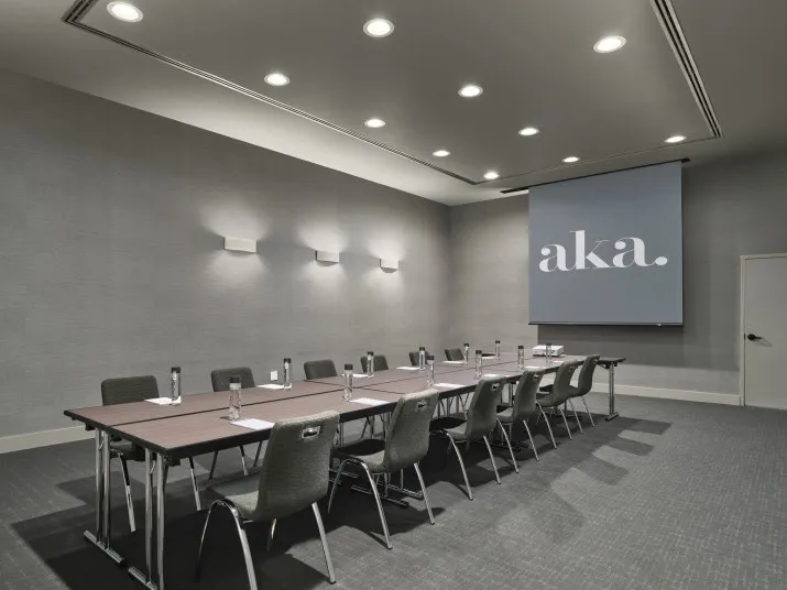 A modern conference room with a long table, several chairs, notepads, pens, and water glasses. A large screen at the front displays the word aka. The room has grey walls and ceiling lights.