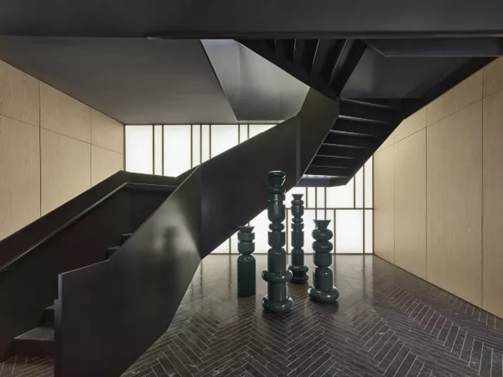 A modern interior with a black spiral staircase, herringbone-patterned floor, light wood-paneled walls, and a cluster of glossy green abstract sculptures at the base of the stairs.