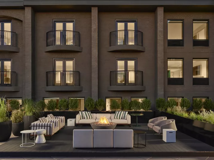 Modern outdoor patio with cushioned seating, striped pillows, a fire pit, potted plants, and a dark brick building in the background featuring balconies and large windows.