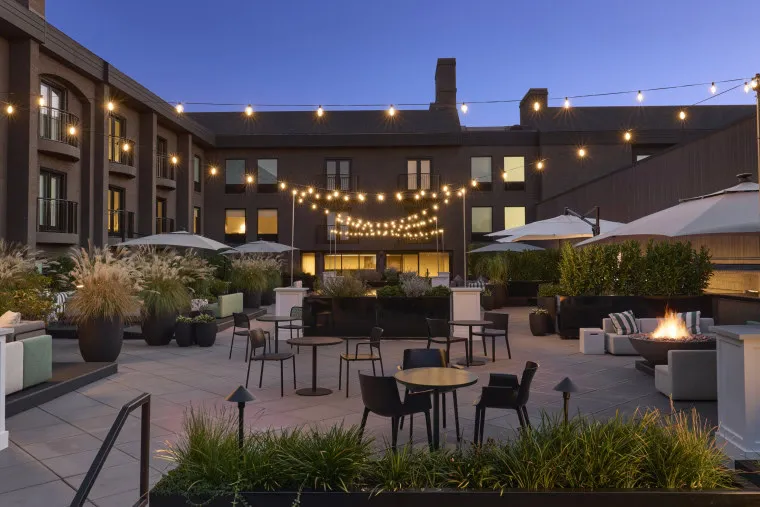 A modern outdoor patio at dusk features string lights, tables and chairs, umbrellas, plants, and a lit fire pit, set against a backdrop of a multi-story building with large windows.