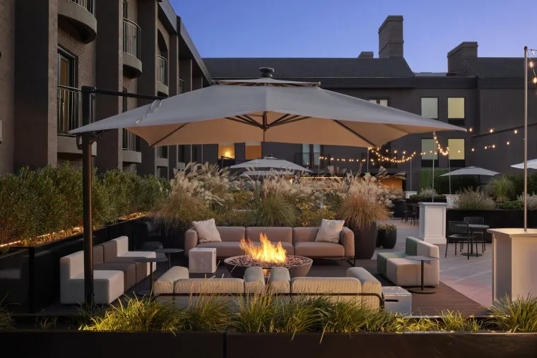 A stylish outdoor patio with modern seating, a round fire pit, large umbrellas, string lights overhead, and tall grasses in planters, set against a backdrop of a building at dusk.