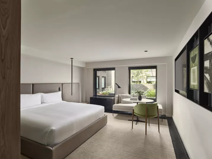 A modern hotel room with a large bed, two windows with garden views, a cushioned bench, a small round table, and a green chair. Neutral tones and minimalist decor create a calm atmosphere.