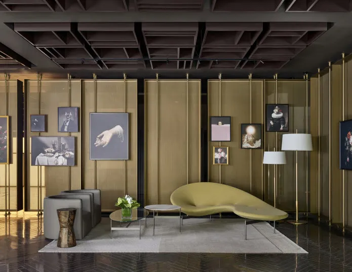 Modern lounge with a sculptural green sofa, gray ottomans, and a small wood side table on a light rug. Framed art hangs on gold mesh walls, and two white floor lamps stand nearby. The ceiling is geometric and dark.