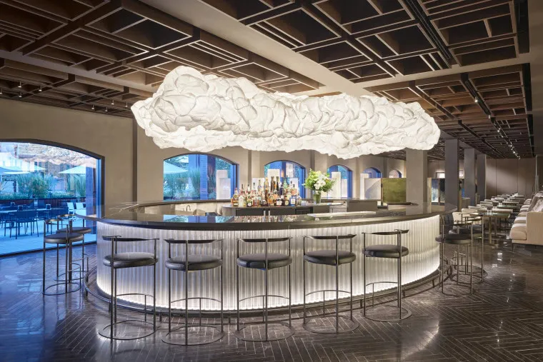 A modern circular bar with high black stools, a glowing white base, and a large cloud-like light fixture above. The space has dark floors, a geometric wood ceiling, and arched windows revealing an outdoor patio.