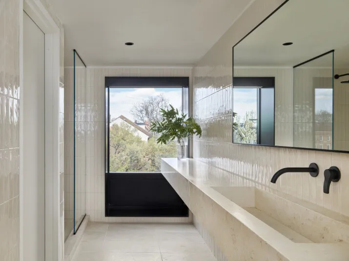 Modern bathroom with beige tiles, a large frameless mirror, a long stone counter with an integrated sink, black wall-mounted faucets, a glass shower, and a large window with outdoor views and a vase of green branches.