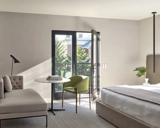 A bright hotel room with a bed, a beige chaise lounge, a small round table, a green chair, and a black lamp. French doors open to a balcony with trees and a building visible outside.