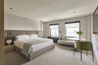 A modern, minimalist hotel room with a large bed, white bedding, two windows, a small sofa, a round table, a green chair, and neutral-colored decor. Natural light fills the clean, spacious space.