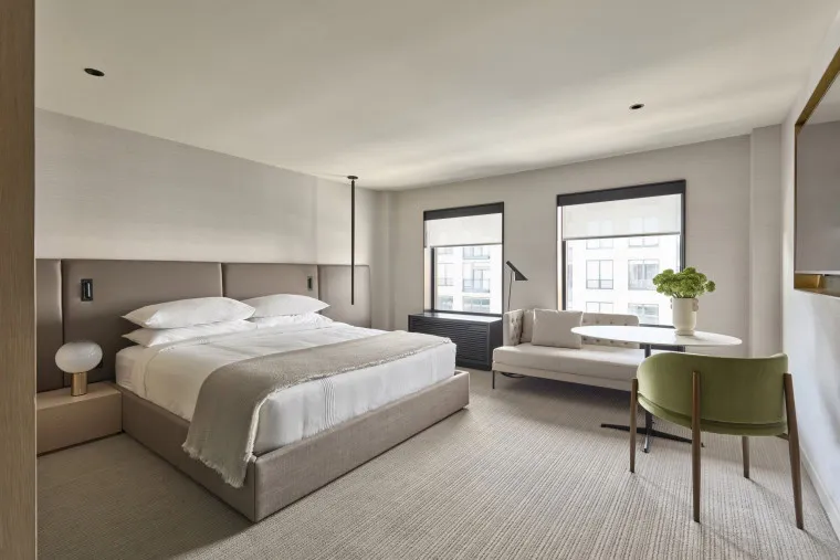 A modern, minimalist hotel room with a large bed, white bedding, two windows, a small sofa, a round table, a green chair, and neutral-colored decor. Natural light fills the clean, spacious space.