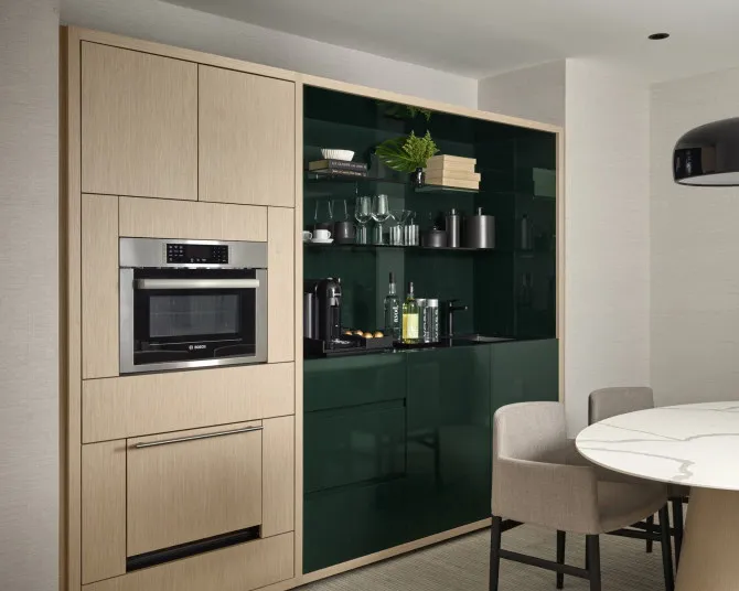 Modern kitchen with light wood cabinetry, dark green glossy cabinets, built-in microwave and oven, open shelves with glassware, coffee machines on the counter, and a round dining table with upholstered chairs.