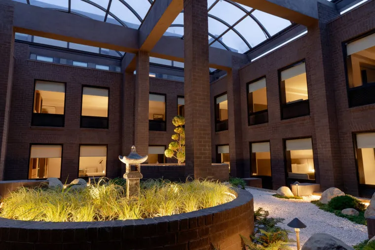 A modern indoor courtyard with a glass roof, brick walls, multiple windows, and a central garden featuring ornamental grasses, a stone lantern, small trees, and soft lighting at dusk.
