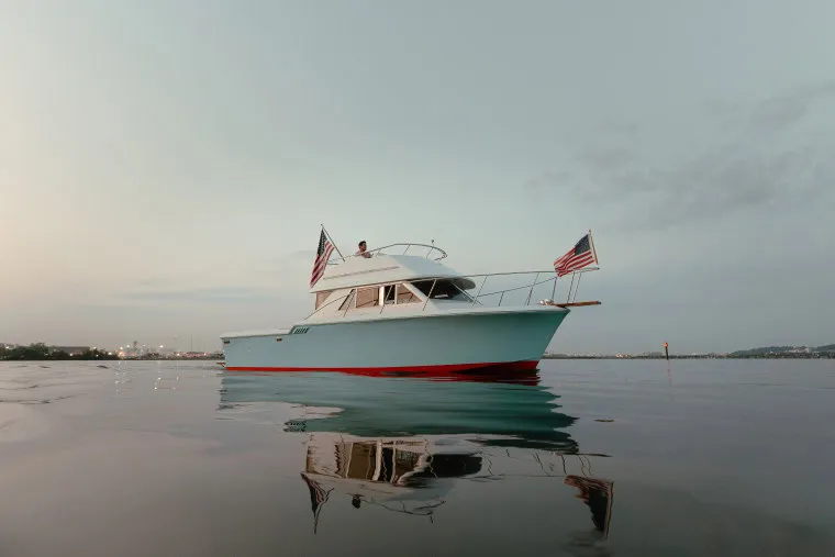 A white and red boat with two American flags is floating on calm water at dusk, reflecting on the surface. The sky is light and clear with a few clouds.