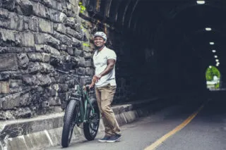 A man wearing a helmet and casual clothes smiles while standing next to a bicycle on a road beside a stone wall, near the entrance of a tunnel.