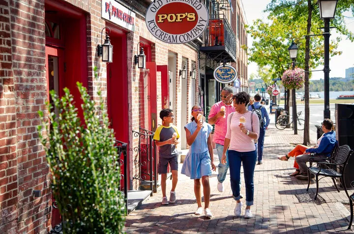 A smiling family enjoys ice cream while walking on a sunny cobblestone sidewalk in front of Pop’s Old Fashioned Ice Cream Company, with other people relaxing on benches nearby.