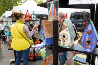 A woman in a yellow vest browses colorful, decorative birdhouses displayed on shelves at a fall outdoor market, with tents and other shoppers visible in the background.