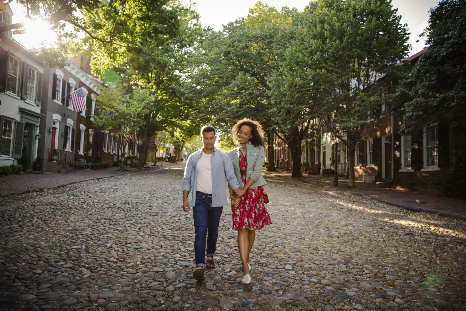 A couple walks hand-in-hand down a cobblestone street lined with trees and historic brick houses during daylight. Sunlight filters through the leaves, creating a warm, peaceful atmosphere.